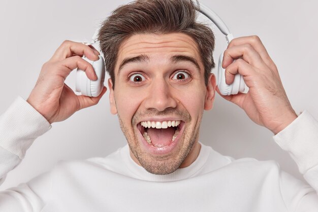 Headshot of happy surprised European man takes off headphones listens music with loud sound from playlist stares amazed at camera exclaims loudly isolated over white background Wow thats great