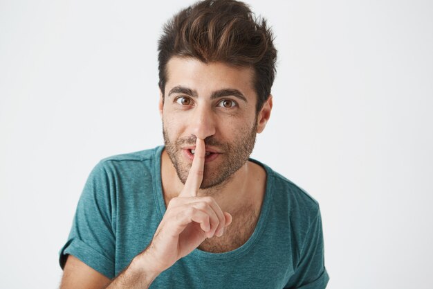 Headshot of happy unshaven smiling hispanic man in casual clothes, holding index finger at lips, asking his girlfriend be quiet after she get extra excited for birthday gift.