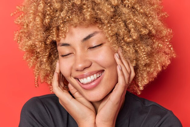 Headshot of happy woman keeps hands on cheeks closes eyes smiles toothily recalls nice memories wears casual clothes has blonde cury hair isolated over vivid red background. Positive emotions