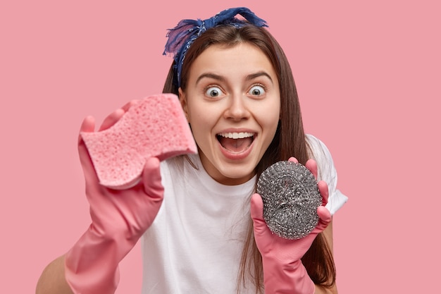 Headshot of happy young woman gazes with joy, has mouth opened, keeps germs away, wipes dust with sponges, wears rubber gloves, headband, finishes household duties, poses over pink wall