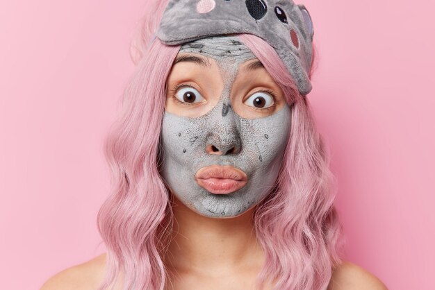 Headshot of impressed young Asian woman stares bugged eyes at camera keeps lips rounded applies clay mask on face for skin treatment wears sleepmask on forehead isolated over pink background