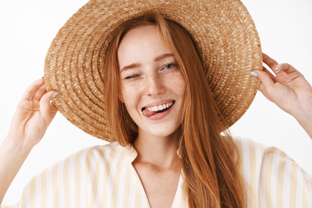 Headshot of joyful attractive redhead female in straw hat with freckles winking happily and sticking out tongue enjoying sunny warm summer day on vacation walking on beach smiling broadly