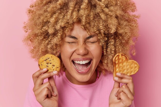 Headshot of joyful European woman with curly hair holds delicious waffle and cookie enjoys eating homemade desserts exclaims from happiness wears casual t shirt isolated over pink background