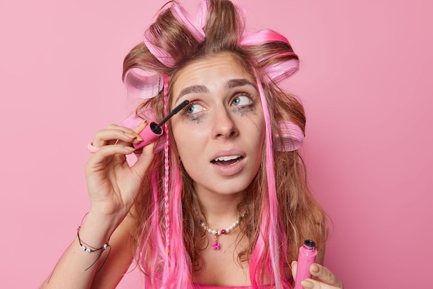 Headshot of long haired young European woman applies hair rollers and mascara looks away undergoes beauty treatments prepares for first date wants to look beautiful isolated over pink background