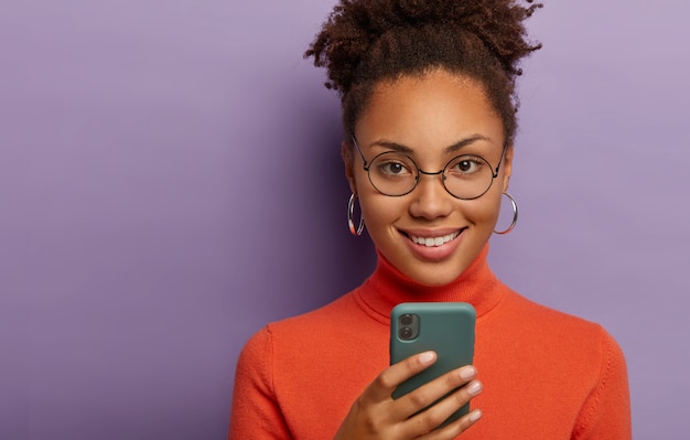 Headshot of lovely curly dark skinned woman uses mobile phone, smiles pleasantly, wears round spectacles, orange jumper, isolated on purple wall. Technology, chatting concept
