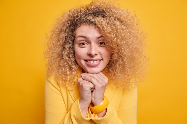 Headshot of lovely curly haired woman keeps hands under chin smiles gently being in good mood 