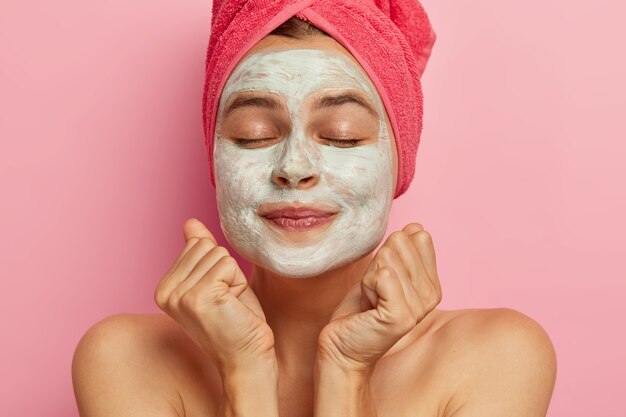 Headshot of lovely girl keeps clenched fists near face, wears facial cleansing mask, keeps eyes shut, has bare shoulders, healthy perfect skin, cares about her body