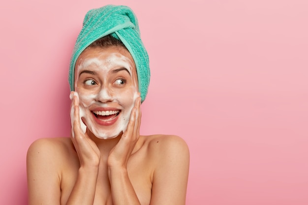 Headshot of lovely smiling female model touches cheeks, looks aside, washes face with soap bubble, has naked body, wears turquoise soft towel on head, poses over rosy wall, copy space for promotion