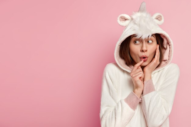 Headshot of lovely surprised cute young woman looks away, has widely opened eyes, wears soft kigurumi costume with hoody, isolated over pink wall, free space for your promotional content.