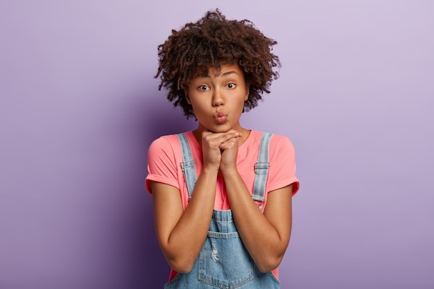 Foto gratuita il primo piano del volto di una donna adorabile tiene le mani sotto il mento, ha le labbra piegate, una pelle sana e scura, un'acconciatura afro