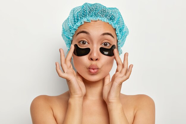 Headshot of lovely woman wears bathcap, applies under eye patch mask for soothing and renewing, keeps lips folded, reduces fine lines, stands with bare shoulders indoor against white wall