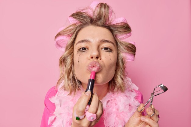 素敵な若い女性のヘッドショットは、口紅を適用し、プロのメイクアップを作るためにまつげカーラーを保持しますピンクの背景に対してヘアローラースタンドに化粧品を使用します朝のルーチン