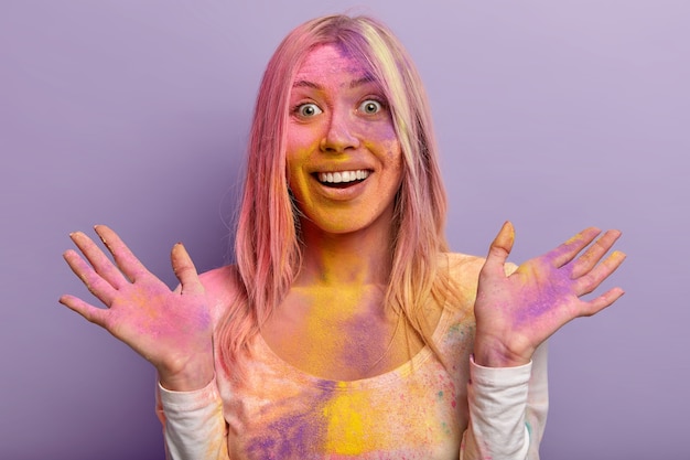 無料写真 歯を見せる笑顔、幸せな反応、手を広げ、色とりどりの乾燥粉末で汚れた嬉しい女性のヘッドショットは、紫色の壁から隔離された伝統的なホーリー祭を楽しんでいます。鮮やかな色