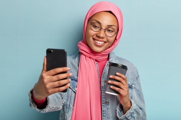 Headshot of optimistic cheerful female wears spectacles, pink veil, tilts head and gazes happily at camera of smart phone, drinks takeaway coffee