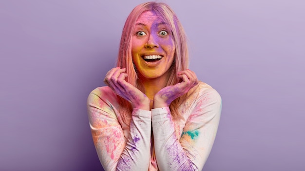 Headshot of optimistic cheerful woman has broad smile, keeps hands near face, has colorful face soiled with holi paints, prepares for spring festival with satisfaction and joy