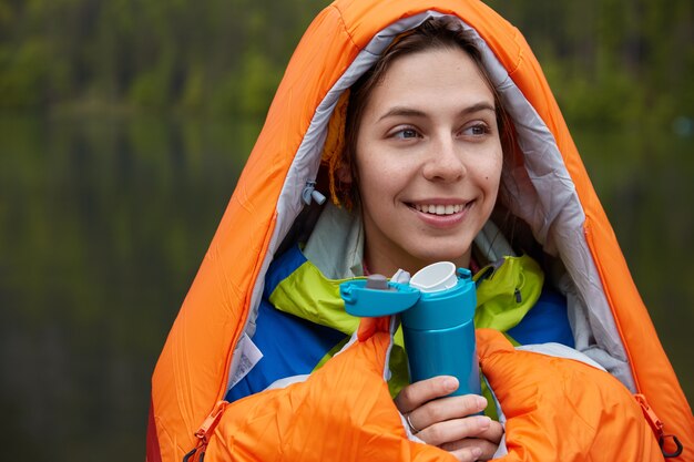 Headshot of optimistic European young woman drinks hot beverage, holds flask, wrapped in orange sleeping bag