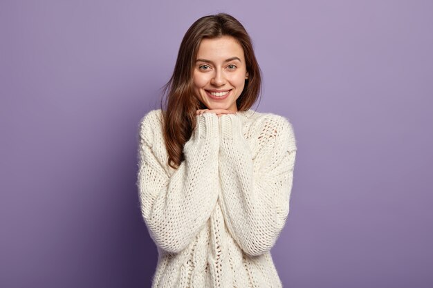 Headshot of optimistic lovely woman with straight hair, keeps hands under chin, wears white jumper with happiness, poses against purple wall People and emotions concept