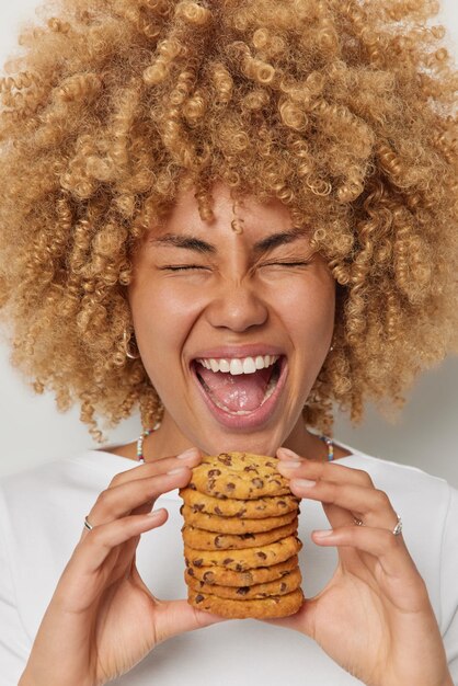headshot of optimistic woman holds pile of delicious cookies with chocolate exclaims and feels very happy keeps eyes closed mouth widely opened dressed casually tasty dessert and nutrition