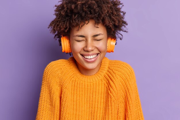 Headshot of overjoyed curly dark skinned teenage girl laughs positively closes eyes enjoys loud sound and favorite song in headphones wears casual jumper spends free time on listening music.