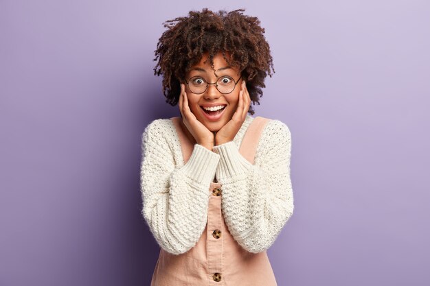 Headshot of overjoyed glad Afro American dark skinned female touches cheeks with both hands, looks with happiness, surprised and happy to receive wonderful present, isolated over purple wall