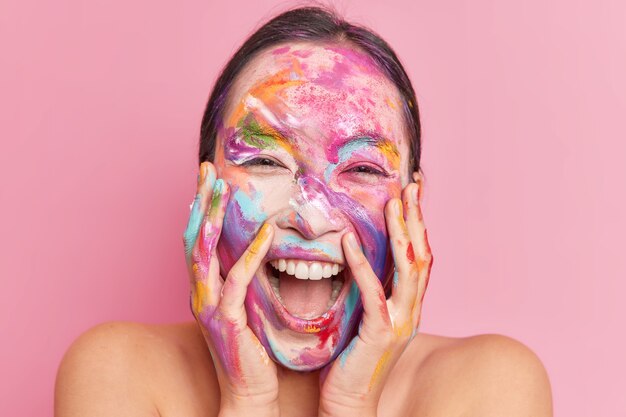 Headshot of overjoyed happy ethnic woman keeps hands on cheeks giggles positively keeps mouth opened has creative makeup smeared face with watercolor paints