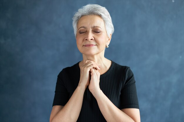 Headshot of peaceful beautiful mature female with short gray hair posing isolated having hopeful facial expression, keeping eyes closed, holding hands clasped in prayer, hoping for the best