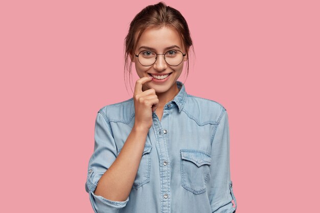 Headshot of pleasant looking friendly woman keeps index finger near mouth, smiles positively