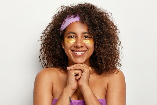 Headshot of pleasant looking lady with toothy smile, Afro bushy hair, stands half naked, keeps hands together under chin, applies yellow patches under eyes for skin recovery