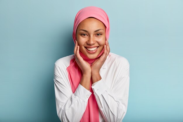 Headshot of pleasant looking Muslim woman touches cheeks with both hands, shows white teeth, wears white shirt and pink veil, isolated against blue wall, expresses joy, happiness, delight