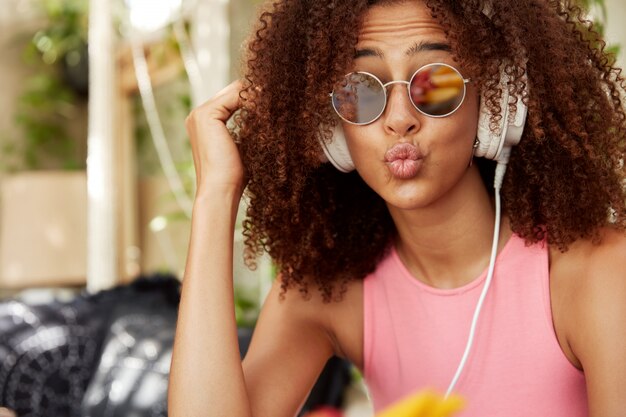 Headshot of pleasant looking styish female in shades has African hairstyle, rounds lips, has funny expression, enjoys favourite music or audio in headphones on radio website. People and style concept