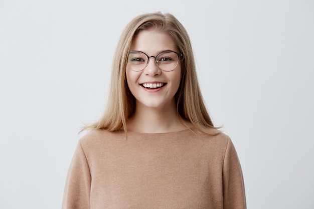 Free photo headshot of pleasant-looking young caucasian woman wearing eyeglasses with broad smile showing her straight white teeth being happy because of positive news. blonde girl with pleasant smile