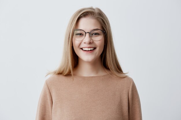 Headshot of pleasant-looking young Caucasian woman wearing eyeglasses with broad smile showing her straight white teeth being happy because of positive news. Blonde girl with pleasant smile