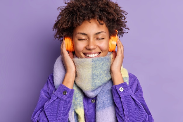 Free photo headshot of pleased curly beautiful young woman closes eyes enjoys favorite melody keeps hands on wireless headphones wears scarf purple jacket.