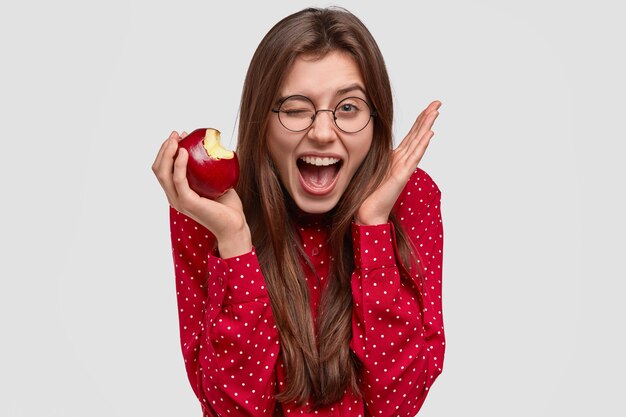 Headshot of pleased young woman blinks eyes, raises hand near head, bites fresh apple, has joyful expression, dressed in red polka dot blouse