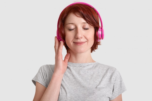Headshot довольной молодой женщины носит наушники, слушает любимую музыку, закрывает глаза от удовольствия, наслаждается громким звуком, одетый в повседневную футболку, изолированную по белой стене. Концепция хобби