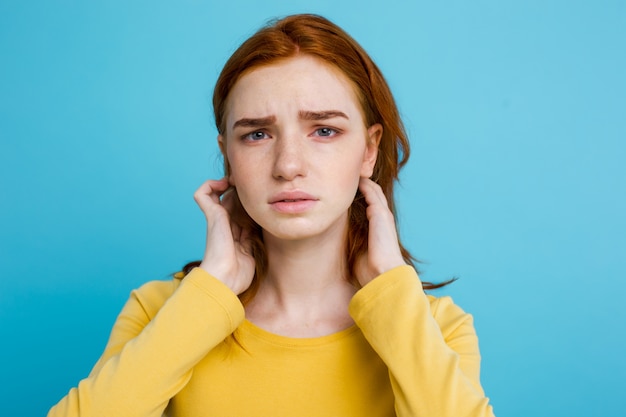 顔を照らす赤面の10代の少女の真剣な表情でカメラを見ている肖像画。屋内でポーズを取る生姜の髪を持つ白人の女性モデル。パステルの青い背景。スペースをコピーします。