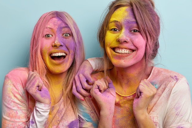 Headshot of positive cheerful two women with toothy smile on faces, have dirty colored skin, raise hands clenched in fists