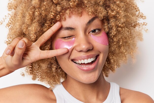 Headshot of positive curly haired woman smiles broadly winks eyes shows peace sign undergoes beauty procedures applies hydrogel pads under eyes isolated over white background. Skin care concept