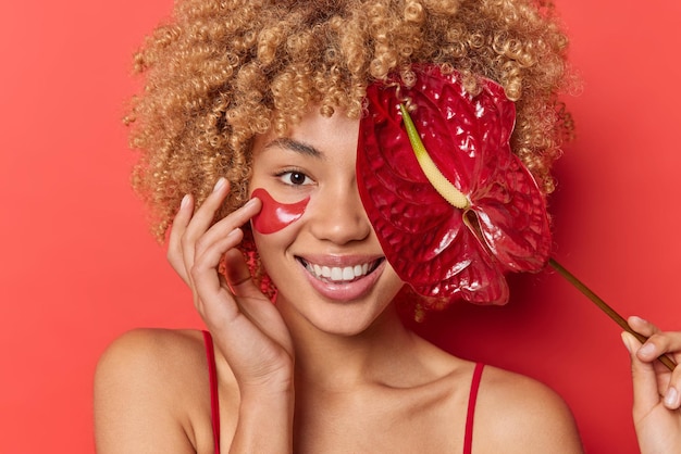 Headshot of positive cute young woman uses organic cosmetic products applies collagen pads and covers eye with calla smiles toothily isolated over vivid red background. Feminine beauty concept