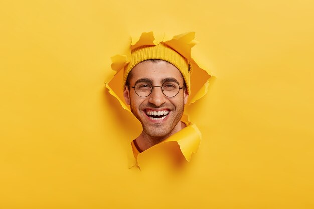 Headshot of positive unshaven young man smiles broadly, wears round optical glasses, yellow headgear