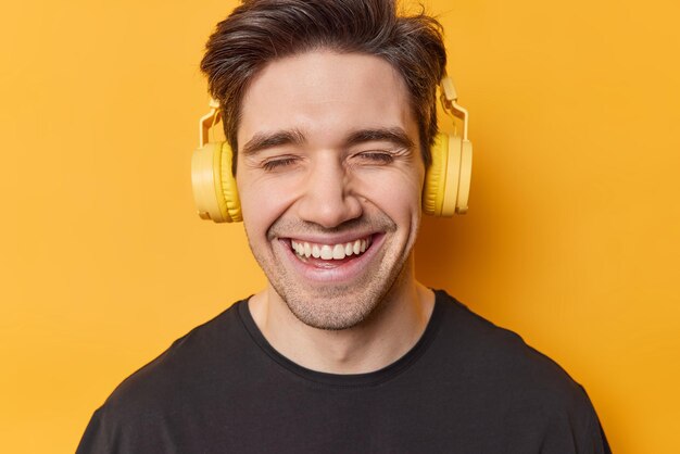 Headshot of positive young man with dark hair enjoys listening music via headphones keeps eyes closed smiles broadly wears black t shirt isolated over yellow background People and hobby concept