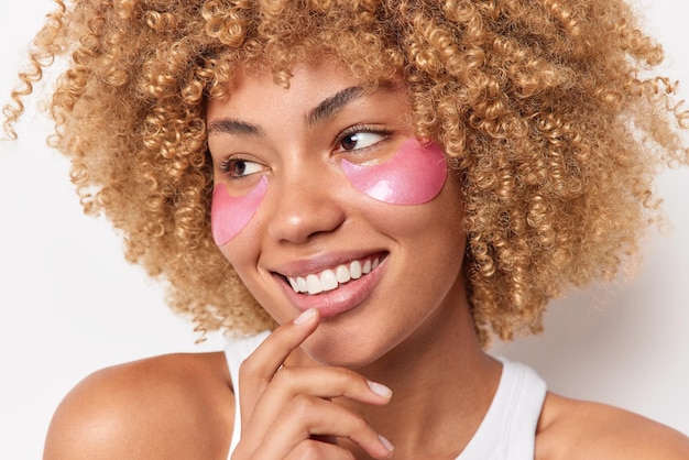 Headshot of positive young woman with curly bushy hair looks away smiles happily applies pink hydrogel patches under eyes isolated over white background. facial treatments and skin care concept