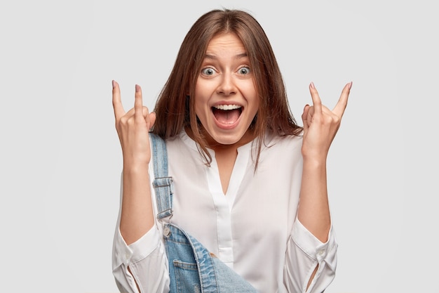 Headshot of pretty cool hipster makes rock and roll gesture, exclaims with happiness, wears stylish outfit