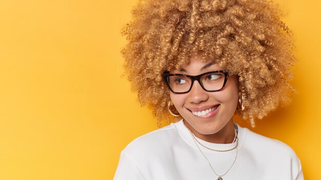 Headshot of pretty curly haired young woman bites lips looks away wih interest wears transparent glasses and white jumper poses against yellow background blank space for your promotional content