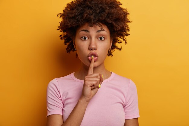 Headshot of pretty curly woman tells secret, hushes to lower voice, asks be speechless, looks directly , presses index finger to lips, wears casual t shirt, isolated on yellow wall