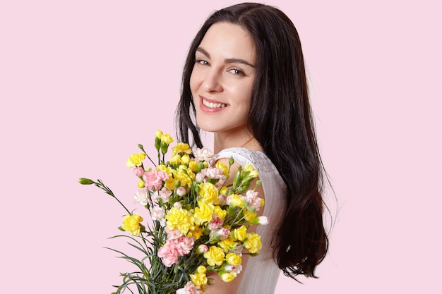 Headshot of pretty European young woman with tender smile, healthy skin, dark long hair, carries bouquet of spring flowers