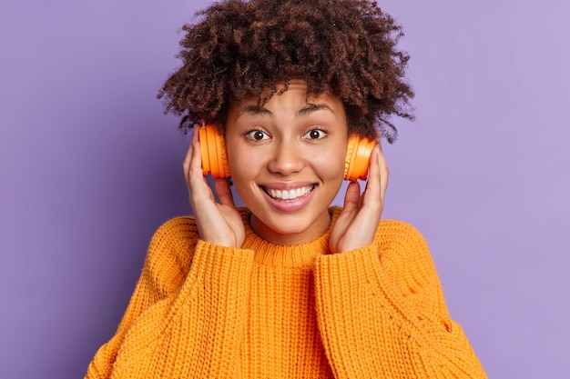 Headshot of pretty joyful African American girl with curly hair keeps hands on headphones smiles toothily wears knitted sweater poses 