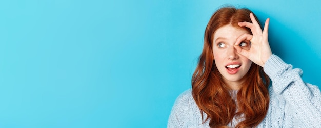 Headshot of pretty redhead girl in sweater looking left at promo with okay sign over eye standing ag
