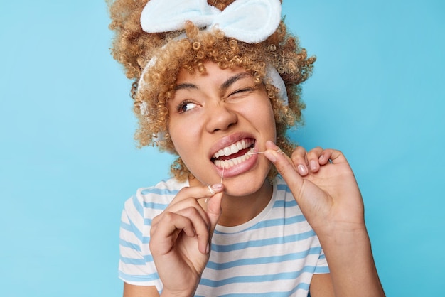 きれいな女性のヘッドショットはデンタルフロスで歯をきれいにします毎日の衛生ルーチンを経てウィンク目は青い壁の上に隔離されたカジュアルな縞模様のTシャツを着て食べた後に食べ物を取り除きますデンタルケア