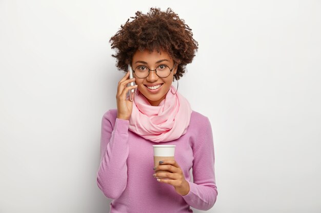 Headshot of pretty young woman with crisp dark hair, enjoys pleasant telephone talk, holds takeaway coffee cup, wears round spectacles, purple jumper, being talkative, uses modern technologies